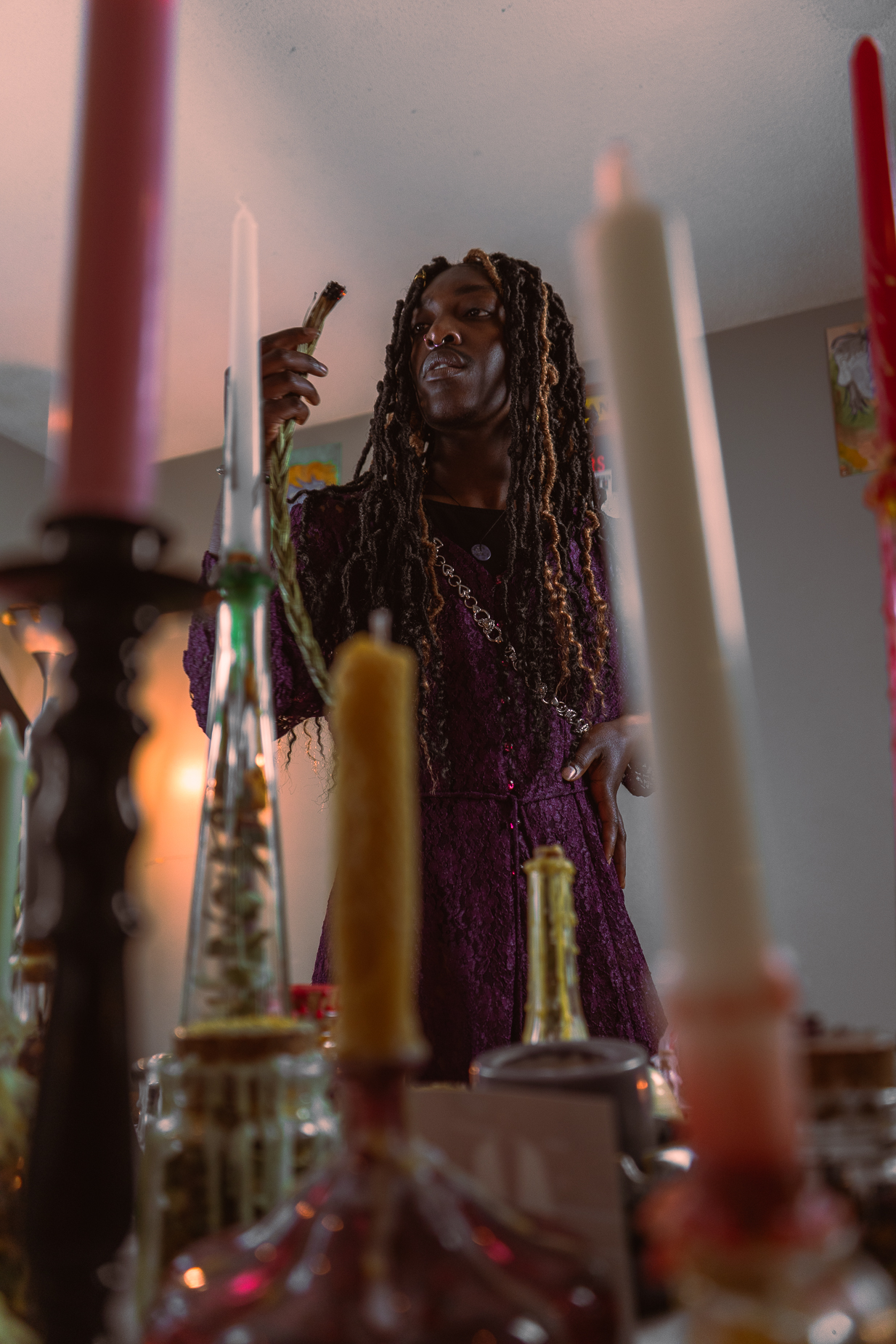 Picture of Viktor Andi-Joi Gee, a young black person with dreads, in their workspace, surrounded by candles, herbs and holding a smudge stick