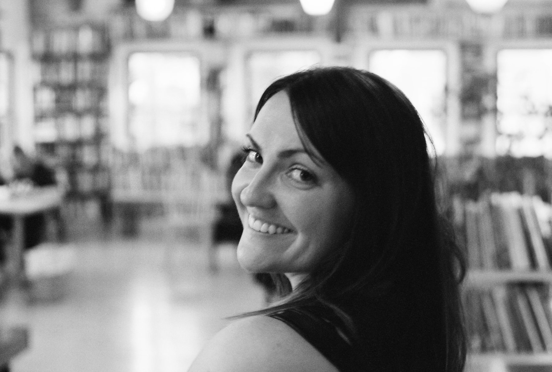 A black and white photo of the head and shoulders of a white woman with dark hair, looking back over her shoulder and smiling