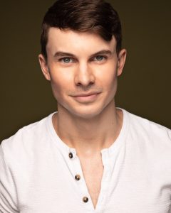 A headshot of a young white man with short dark hair in a loose white open-necked shirt