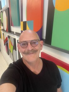 A photo of Kansas CIty artist Kevin Neen - a head shot of a bald white man with a mustache and glasses, wearing a black tshirt against a background of brightly coloured art