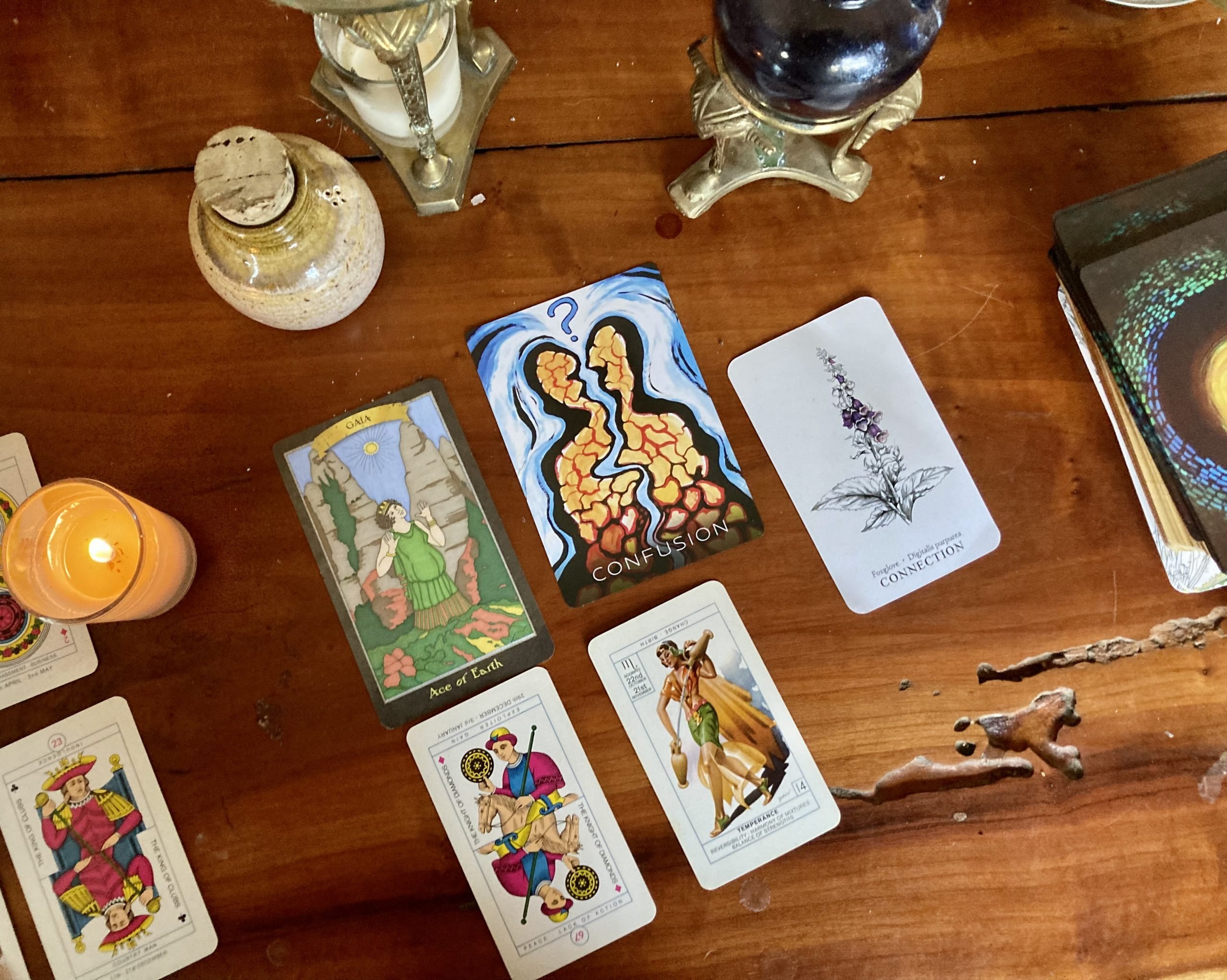 A spread from a several modern tarot decks is visible on a wood table with jars and candles nearby. The cards have different sizes and art styles. Visible are the king of clubs, the knight of diamonds, the ace of earth, temperance, confusion, and connection. The rainbow mosaic pattern of the back of the rest of the cards is also visible.
