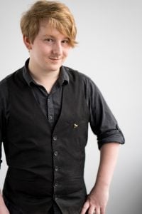 Aaron S. Ricucci-Hill - a photo of a young white person with sandy reddish hair wearing a black shirt and waistcoat, with their sleeves rolled up, standing with their hands on their hips